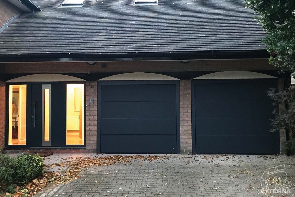 Divi antracīta garāžas vārti un apgaismotas ārdurvis ķieģeļu mājā vakarā